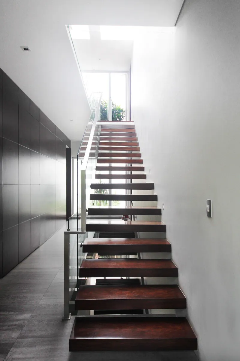 Escalera de peldaños de madera flotantes con baranda de vidrio y luz cenital.